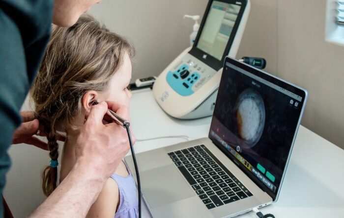 An older man is holding a medical instrument to look into the girl's ears.  In front of this girl, there is a laptop showing the image inside her ear