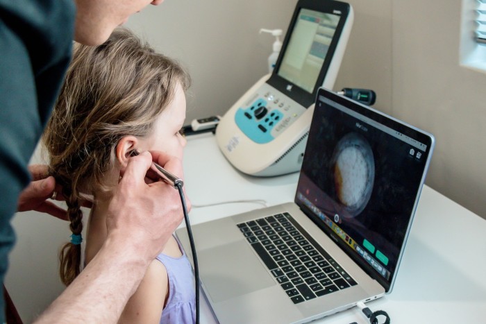 An older man is holding a medical instrument to look into the girl's ears.  In front of this girl, there is a laptop showing the image inside her ear